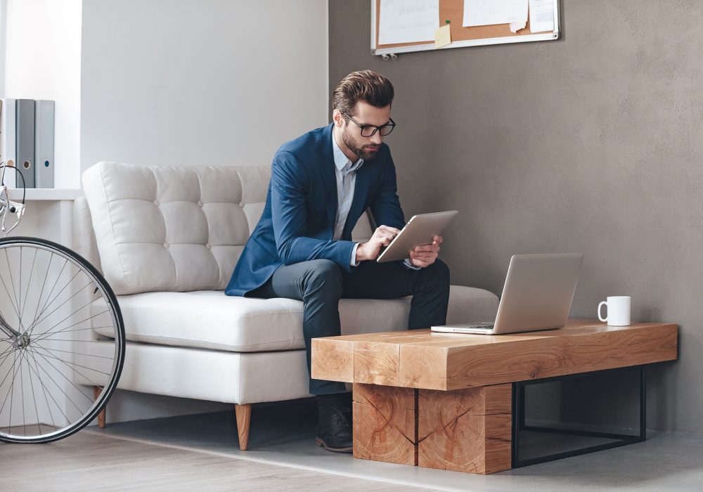 Multitasking. Handsome young man wearing glasses and working with touchpad while sitting on the couch in office Multitasking. Handsome young man wearing glasses and working with touchpad while sitting on the couch in office