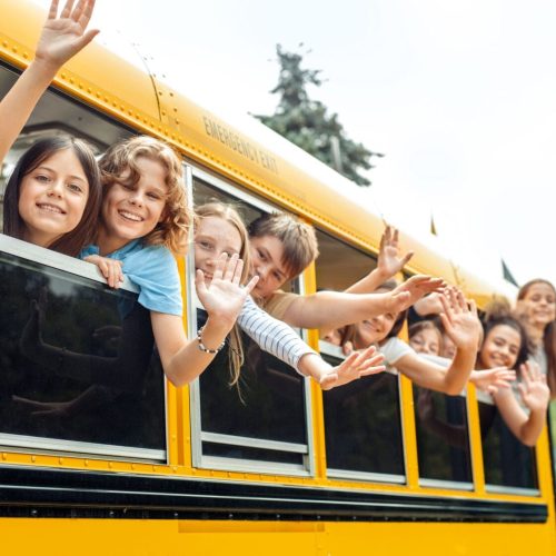 Group of children classmates going to school by bus leaning out of the window waving to camera smiling positive